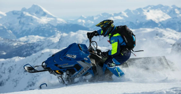 person riding blue snowmobile through fresh snow with mountain range in background